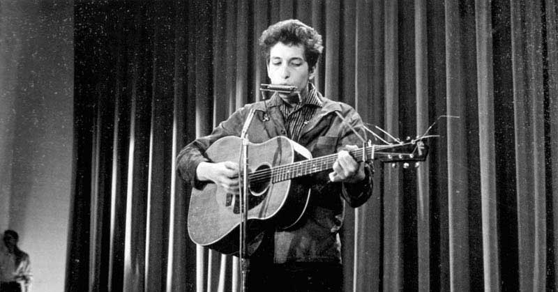 باب دیلن در حال نواختن گیتار و هارمونیکا به صورت همزمان